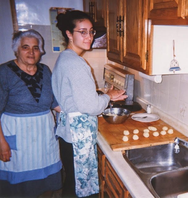 Ariane's nonna stands behind her. They are both wearing aprons, cooking together in the kithcen.