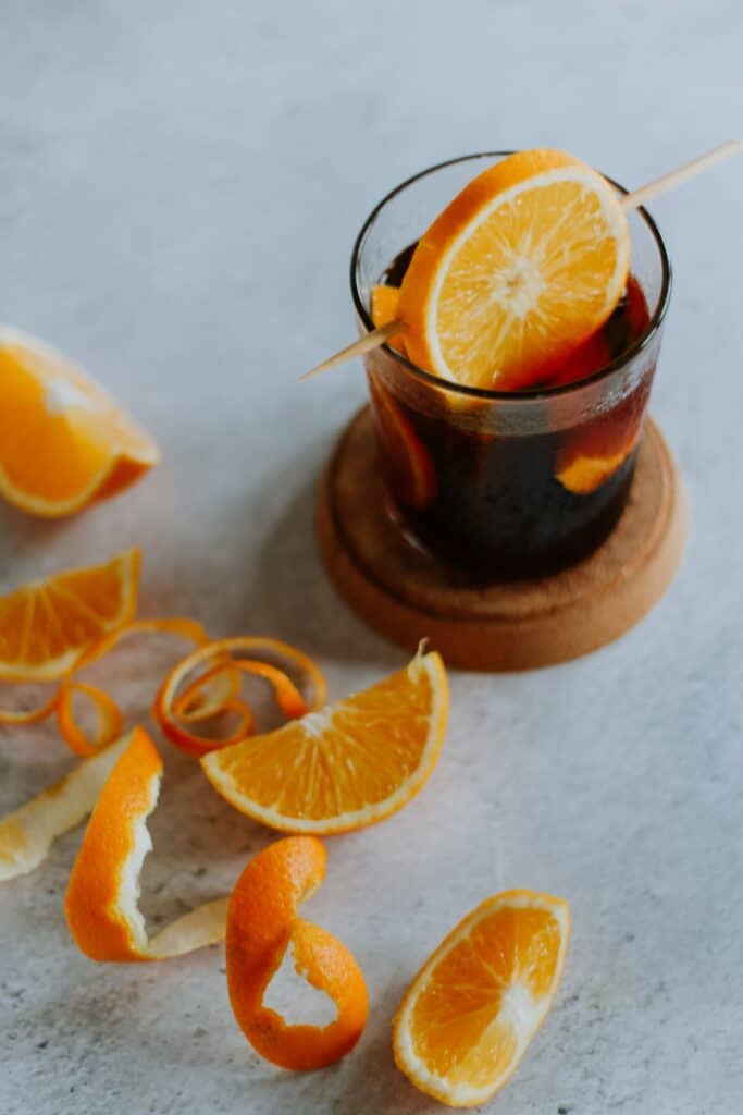 On a light grey table, there is a cocktail with an assortment of oranges and orange peels surrounding it.