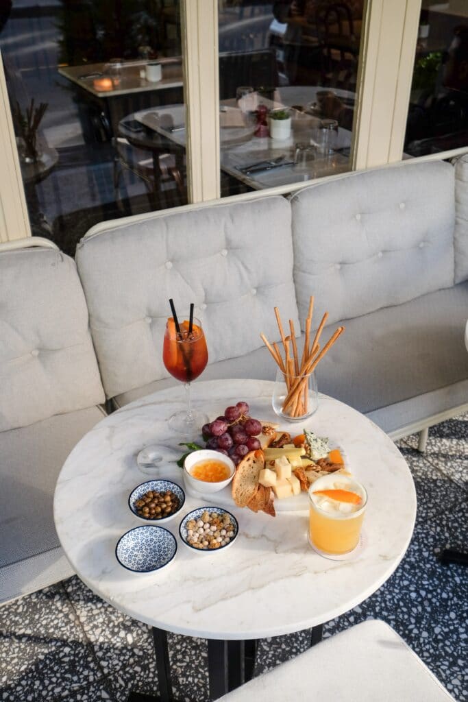 In front of a window, there is a grey sofa. In front of the sofa, there is a white marble table with an assortment of cocktails, fruit, bread and cheeses. The floor is black and white with an abstract pattern.