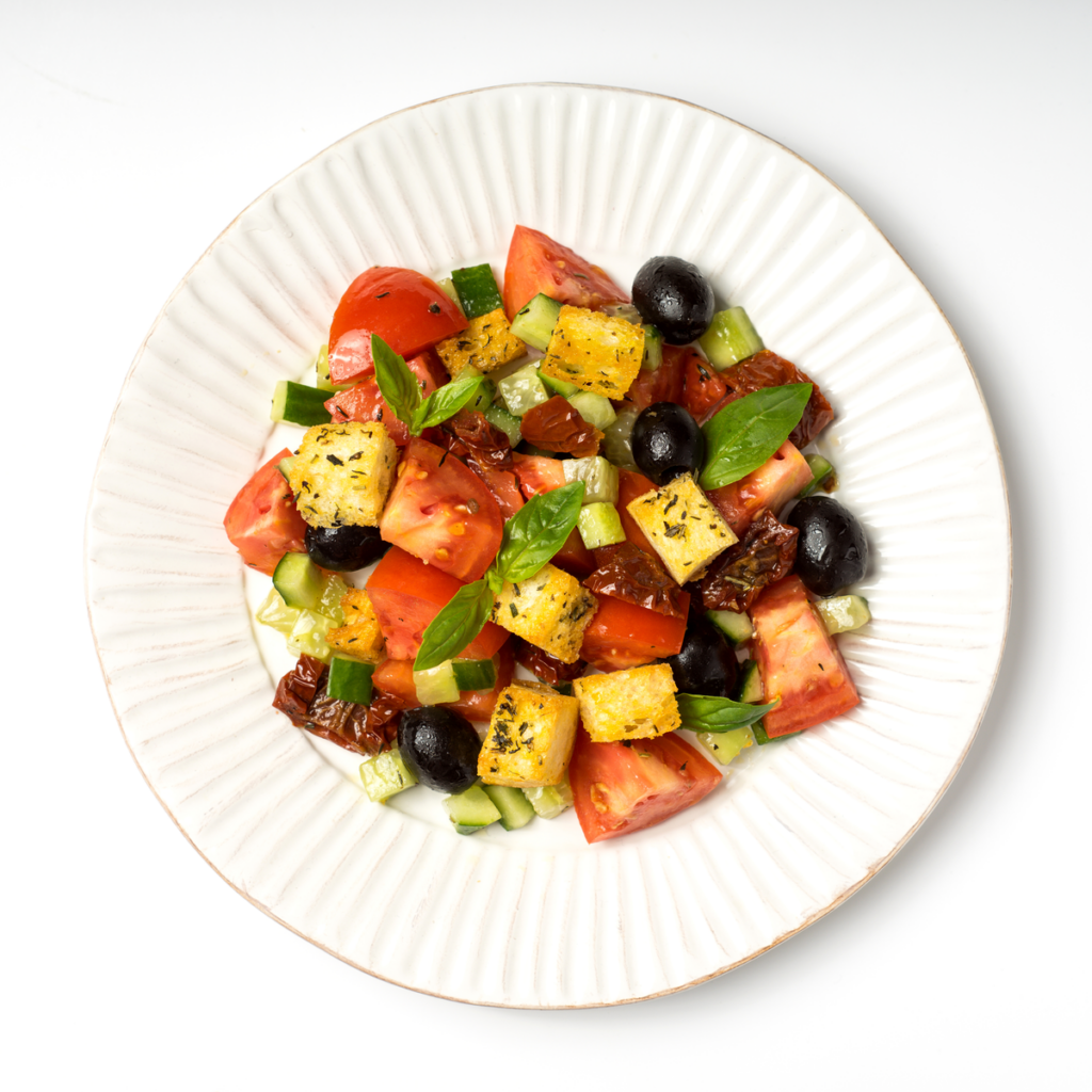 A white plate of Salad that includes basil, tomatoes, black olives, pineapple and cucumbers